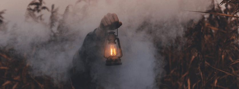 A hand reaches out of the mist carrying an old-fashioned lantern. Only the hand and arm of the person are visible.