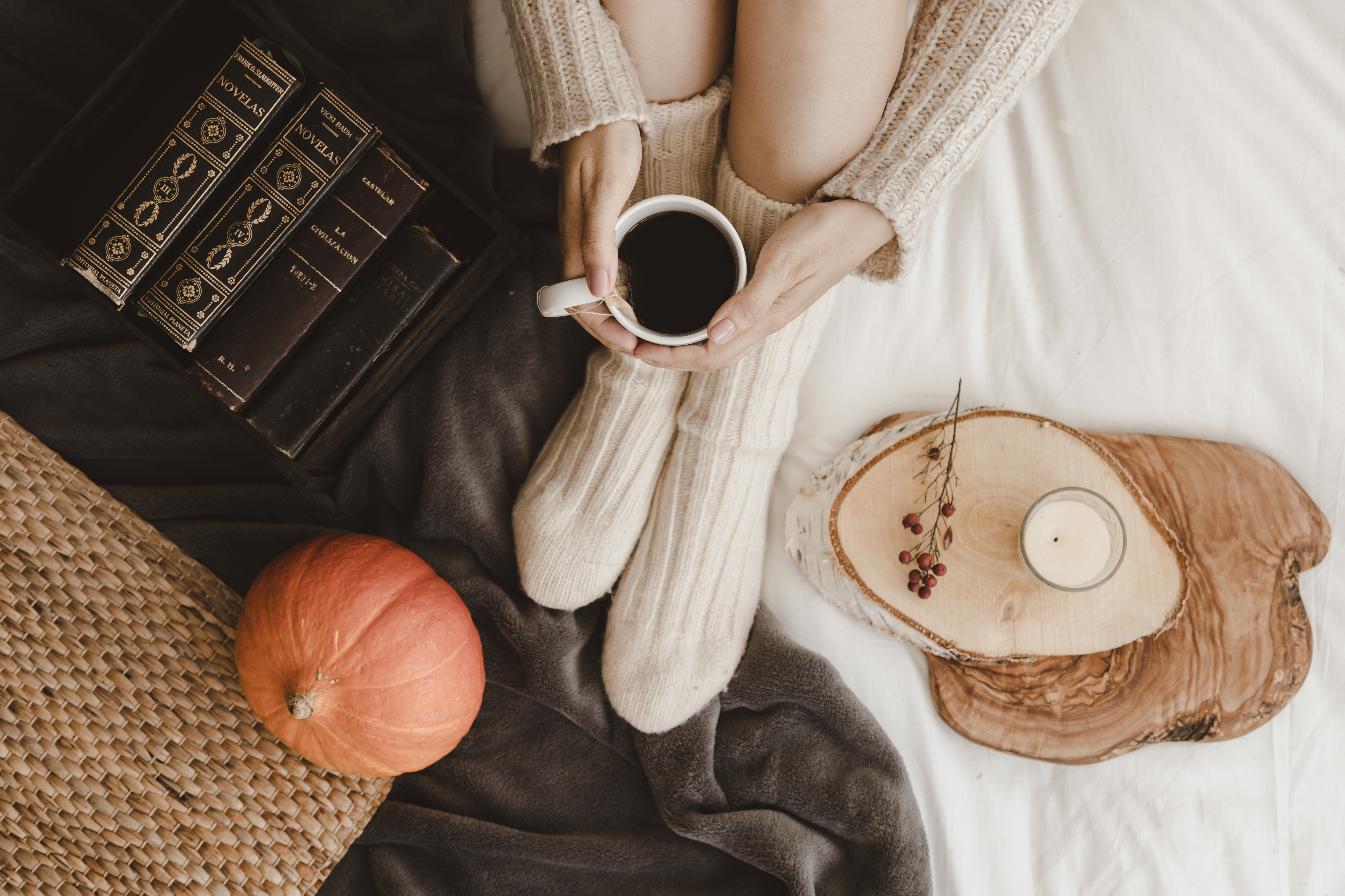 Person with bare legs and knit socks sits on the floor, holding a cup of coffee. They are surrounded by books and autumn decorations.